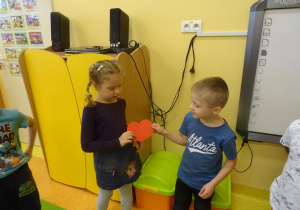 Dwoje dzieci stoi obok siebie, dziewczynka podaje chłopcu papierowe serduszko.